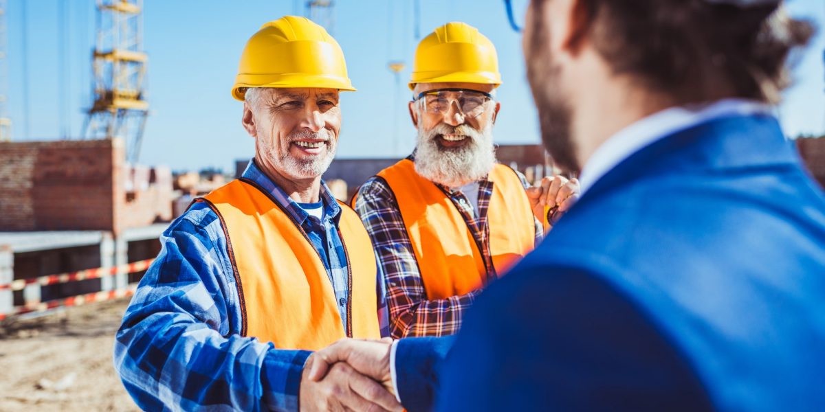 Construction worker in protective uniform shaking hands with businessman in hardhat at construction